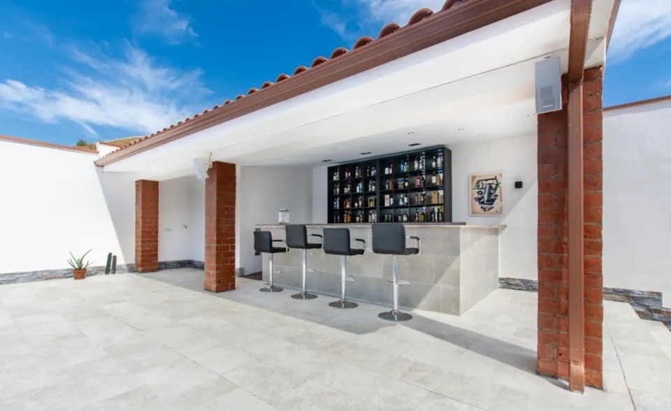 La barra cubierta con sus taburetes y el botellero, junto a la zona de la piscina