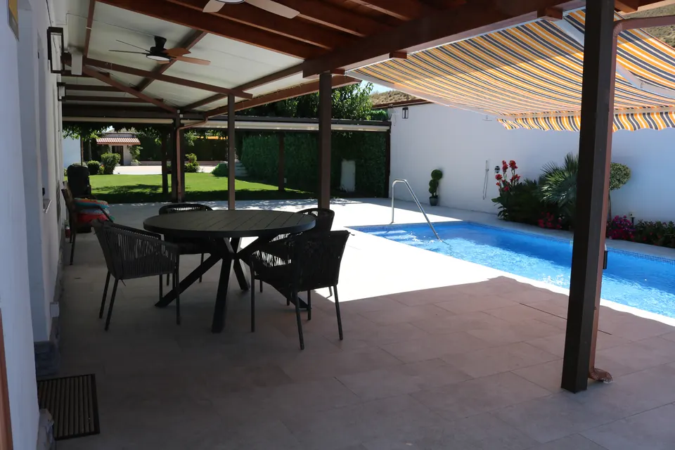 Mesa redonda con sillas a la sombra de la pérgola, con la piscina al lado